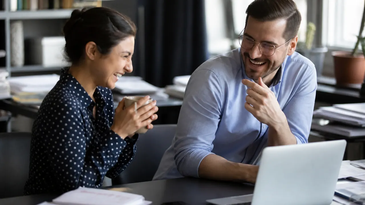 Two coworkers laughing beside a messy desk after a short joke, phone screen not readable
