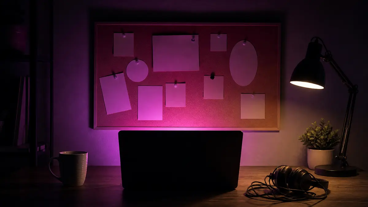 Night studio desk, open laptop facing away with a magenta glow on a corkboard