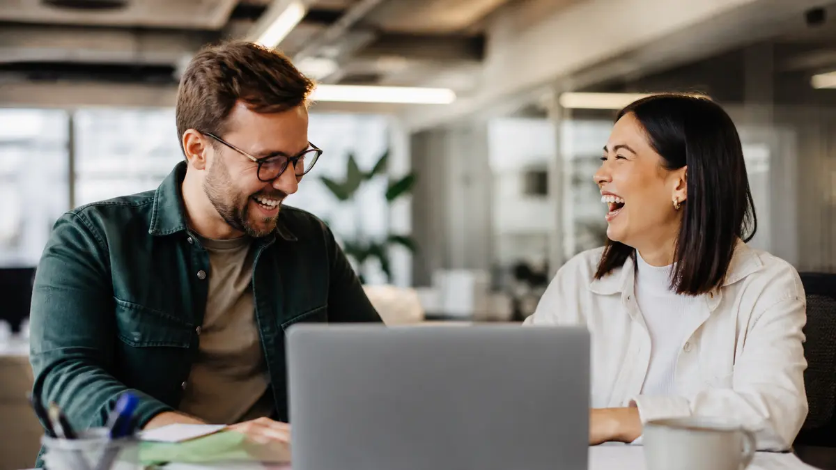 Two coworkers laughing beside a messy desk after a short joke, laptop screen not readable