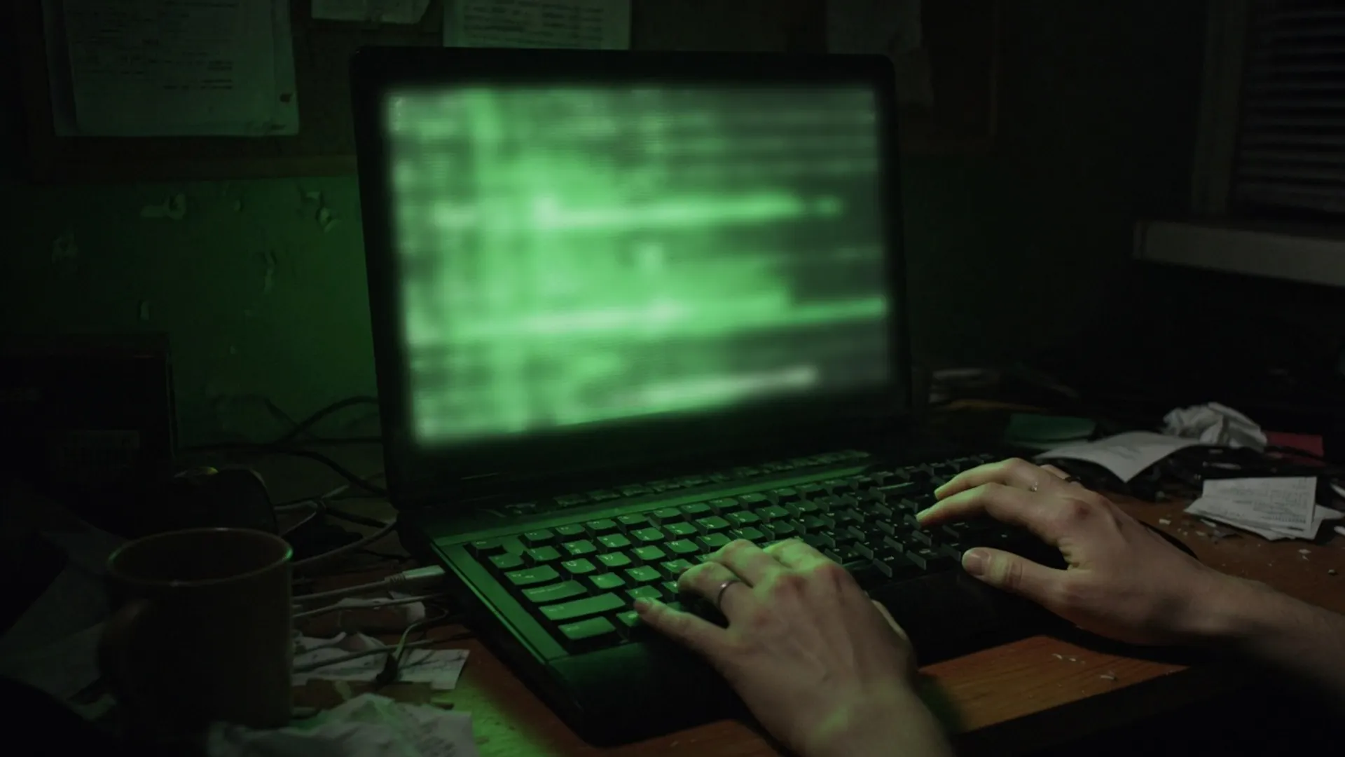 Dark desk with a laptop casting green glow while hands rest on a keyboard, no readable screen text